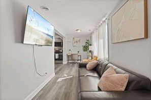 A modern living room with a leather sofa, a flat-screen TV mounted on the wall, a dining area with a table and chairs, and a kitchen in the background. - Modern 4BD 2BA Home Near Downtown Metra (Highland Park)