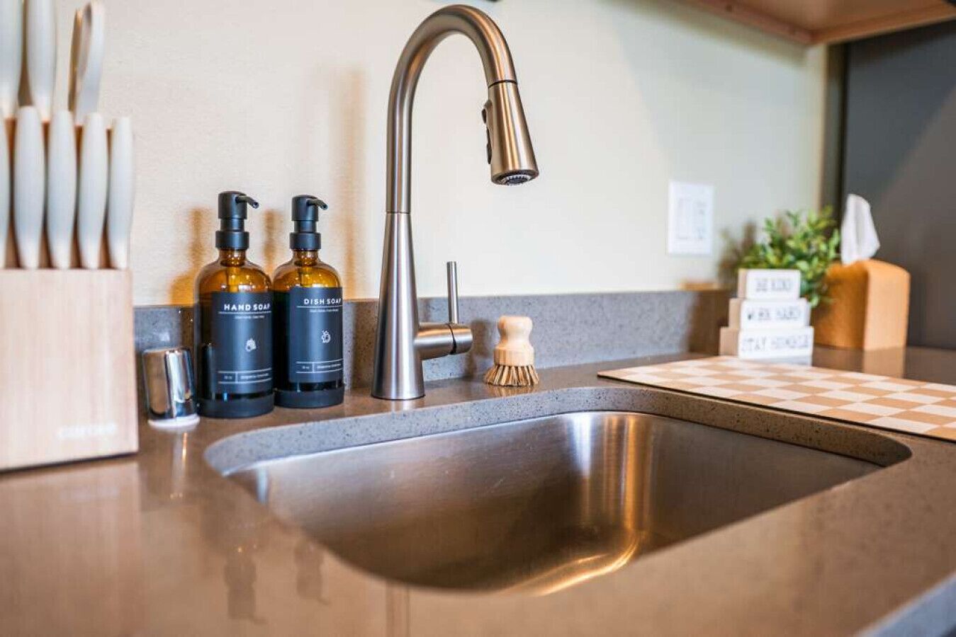 A kitchen sink with a modern faucet, two soap bottles, a soap brush, and a checkered cutting board.