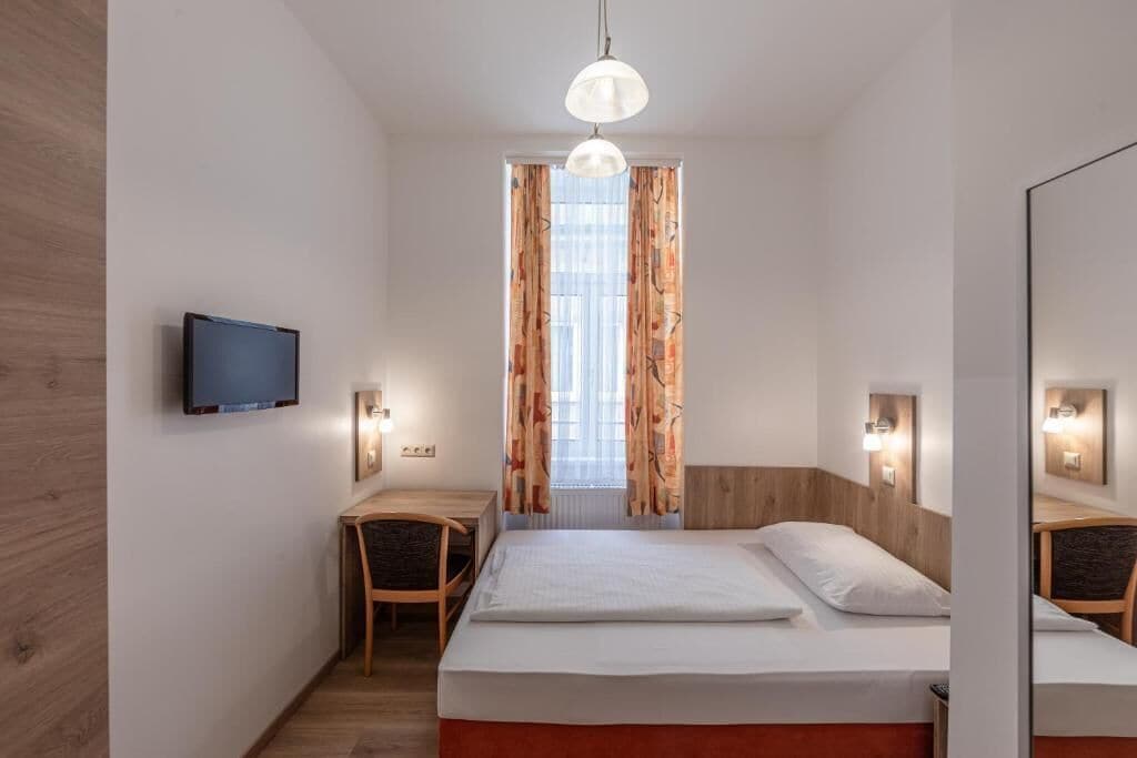A bedroom with a bed, a TV, a desk, and a window with curtains.