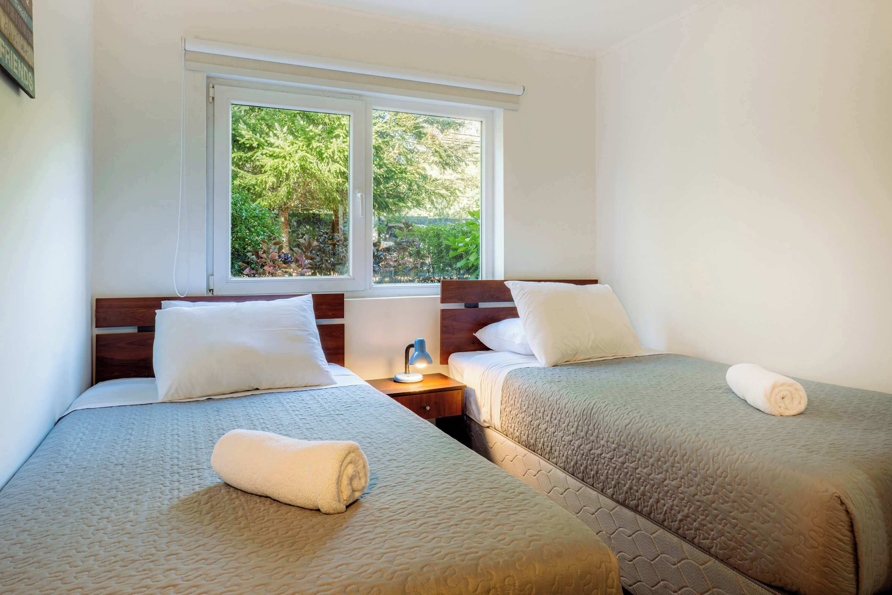 A hotel room with two beds, a window with a view of greenery, a bedside table with a lamp, and a neatly made bed.