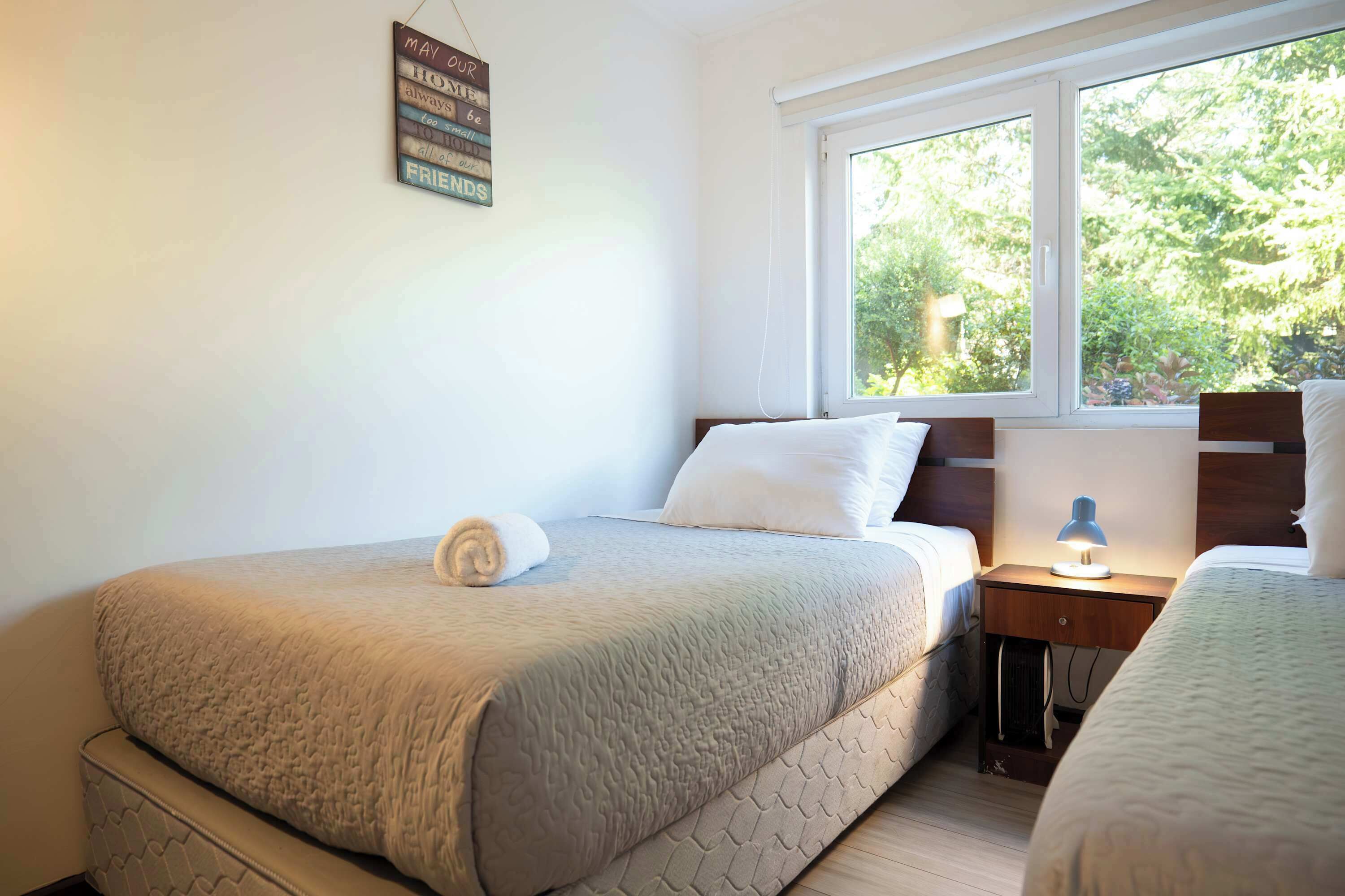 Camera da letto con due letti, una finestra vista alberi, un comodino con una lampada e un cartello appeso al muro.