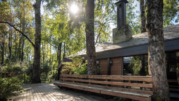 Une terrasse en bois avec un banc, entourée d’arbres et d’un bâtiment doté d’une cheminée.