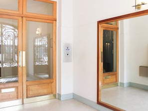 A room with a wooden door, a glass door with a metal gate, a wall-mounted light switch, and a large mirror reflecting part of the room. - Bright balcony near Roland-Garros (Paris)