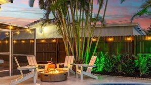 A poolside area with a fire pit, lounge chairs, and a covered seating area. - Toucan Tiki Retreat | Coastal Modern - Heated Pool (Deerfield Beach)