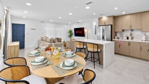A modern kitchen with a central island, a dining area with a round table and chairs, and a living area with a sofa and a TV. - Toucan Tiki Retreat | Coastal Modern - Heated Pool (Deerfield Beach)