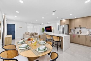 A modern kitchen with a central island, a dining area with a round table and chairs, and a living area with a sofa and a TV. - Toucan Tiki Retreat | Coastal Modern - Heated Pool (Deerfield Beach)