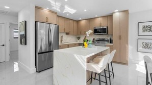 A modern kitchen with a central island, stainless steel appliances, and wooden cabinetry. - Toucan Tiki Retreat | Coastal Modern - Heated Pool (Deerfield Beach)