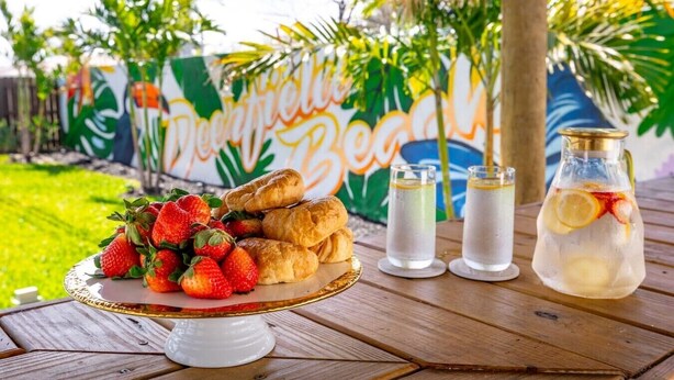 A table with a fruit platter, two glasses of a clear beverage with lemon slices, and a bottle with a similar drink. - Toucan Tiki Retreat | Coastal Modern - Heated Pool (Deerfield Beach)