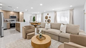 A modern living room with a sofa, chairs, a coffee table, and a kitchen area in the background. - Toucan Tiki Retreat | Coastal Modern - Heated Pool (Deerfield Beach)