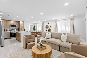 A modern living room with a sofa, chairs, a coffee table, and a kitchen area in the background. - Toucan Tiki Retreat | Coastal Modern - Heated Pool (Deerfield Beach)