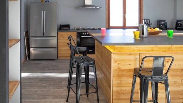 A modern kitchen with a stainless steel refrigerator, wooden bar stools, a wooden island, and a large window.