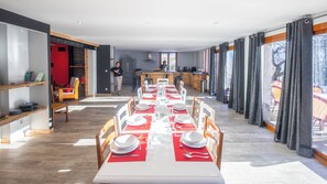 A dining area with a long table set for a meal, a kitchen in the background, and large windows with curtains.