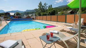 A swimming pool with lounge chairs and umbrellas, surrounded by a wooden fence and mountains in the background.