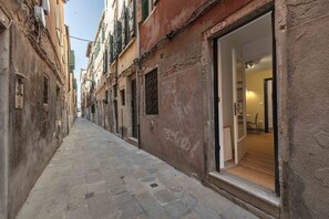 A narrow street with buildings on either side, a visible entrance to a property, and a clear view into a room with a wooden floor and a door. - Daisy Flowers by Wonderful Italy (Venice)