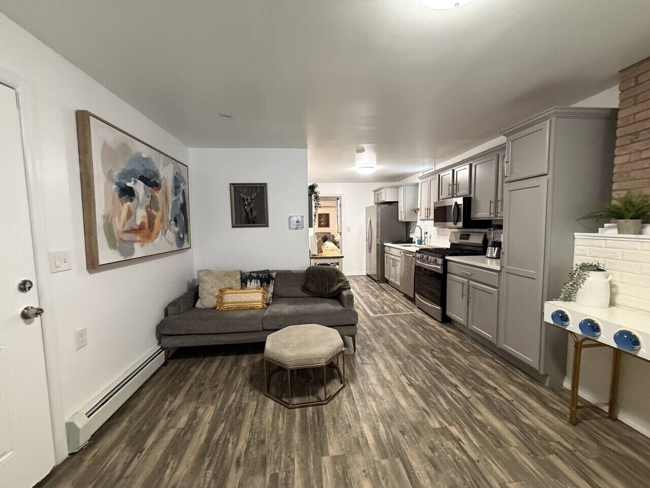 Una cocina moderna con armarios grises, un comedor con sofá y reposapiés grises, y una sala de estar con un cuadro en la pared.