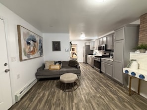 Eine moderne Küche mit grauen Schränken, ein Essbereich mit grauem Sofa und Hocker sowie ein Wohnzimmer mit einem Bild an der Wand.