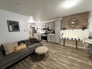 Eine moderne Küche mit weißen Schränken, einer Mikrowelle und einem Herd. Ein graues Sofa mit Decke und ein kleiner Hocker sind ebenfalls vorhanden.
