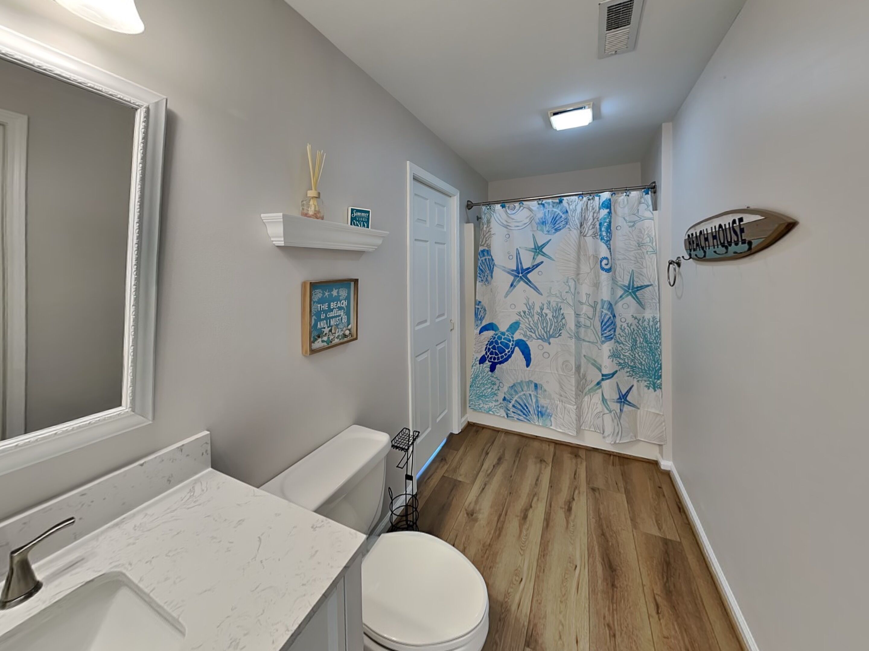 Une salle de bain avec un lavabo blanc, des toilettes, un rideau de douche orné de motifs de tortues de mer, une étagère avec des objets décoratifs et une pancarte indiquant « MAISON DE PLAGE ».