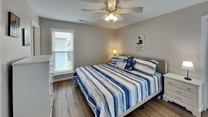 Un dormitorio con cama, ventilador de techo, mesita de noche, cómoda y ventana con persianas.