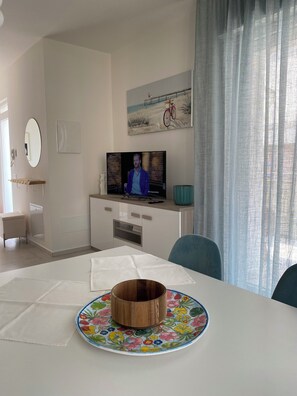 A modern living room with a television, a dining table with a floral plate, a painting of a bicycle, and a window with curtains. - Villa Luigia Garda. "Azure" (Peschiera del Garda)