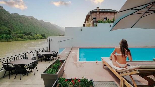 A poolside area with a view of a river, outdoor seating, and a building in the background. - River Front Mtskheta (Mtskheta)