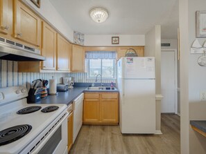 A compact kitchen with wooden cabinets, a white refrigerator, a stove, and a window with blinds. - Sand Pebbles #A3 - Pelican Perch (Carolina Beach)