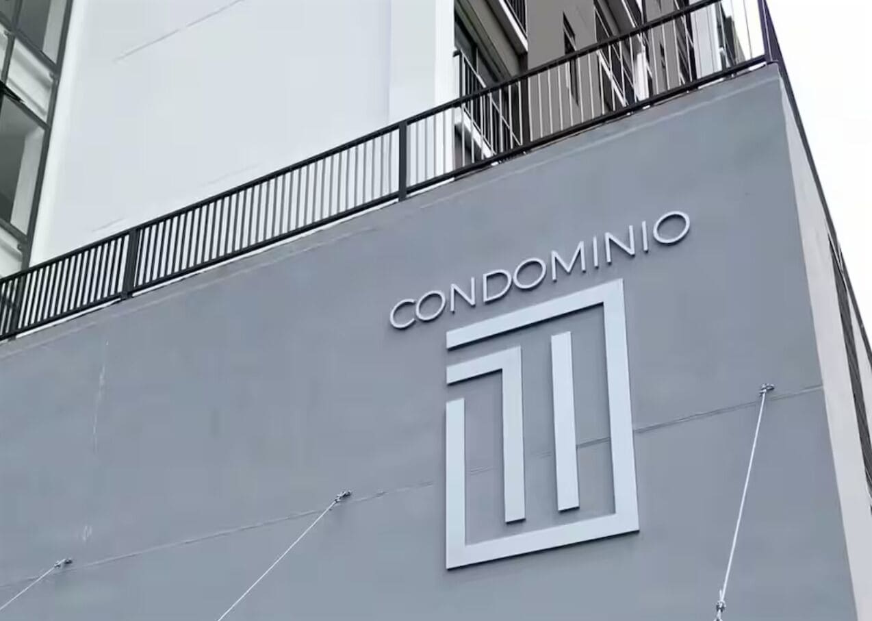 Ein Gebäude mit einer grauen Fassade und einem schwarzen Geländer. Über einem großen, weißen geometrischen Logo ist das Wort 'CONDOMINIO' sichtbar.