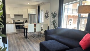 Un salon moderne avec un canapé et un pouf de couleur sombre, un coin repas avec une table et des chaises, une cuisine avec des meubles blancs et des appareils en acier inoxydable, et une grande fenêtre donnant sur un immeuble en briques.