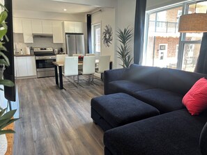 A modern living room with a dark sofa and ottoman, a dining area with a table and chairs, a kitchen with white cabinets and stainless steel appliances, and a large window with a view of a brick building. - Passion, 2 Queen-size bed, Air-conditioned, Balcony, Free parking (Lévis)