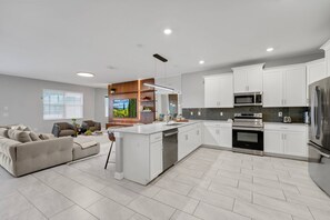 Une cuisine moderne avec des meubles blancs, un îlot central, des appareils en acier inoxydable et un sol carrelé de couleur grise.