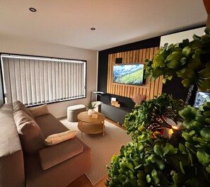 Una sala de estar moderna con un sofá, una mesa de centro, un televisor montado en una pared de madera y un ventanal con persianas.