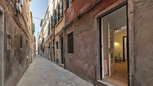 A narrow street with buildings on either side, a visible entrance to a property, and a clear view into a room with a wooden floor and a door. - Daisy Flowers by Wonderful Italy (Venice)