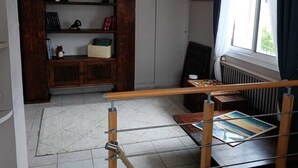 Ett rum med trägolv, ett fönster med metallram och gardiner, ett element och en hylla med böcker och dekorativa föremål.