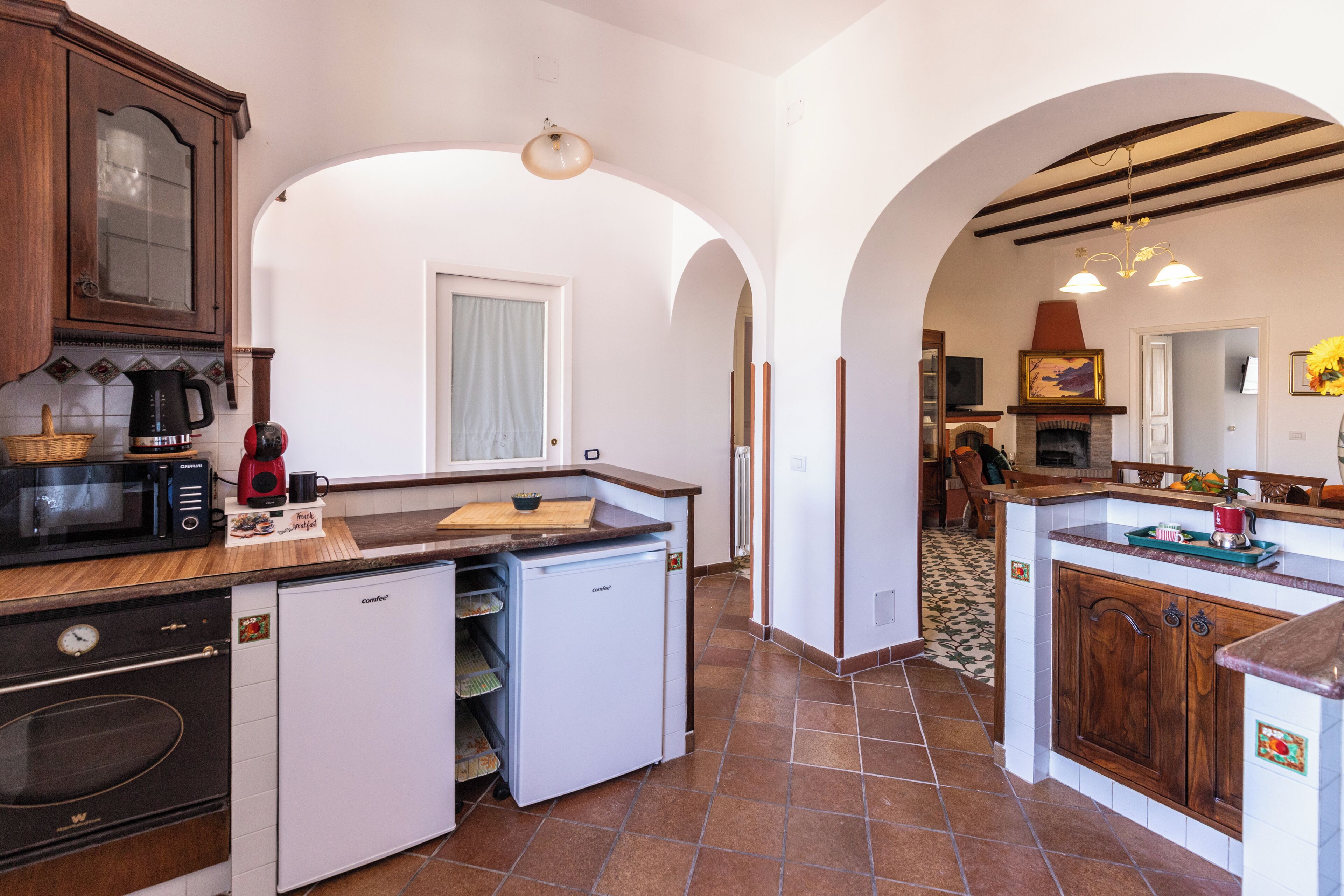 A compact kitchen with a built-in oven, microwave, and refrigerator, an archway leading to a dining area, and a fireplace.