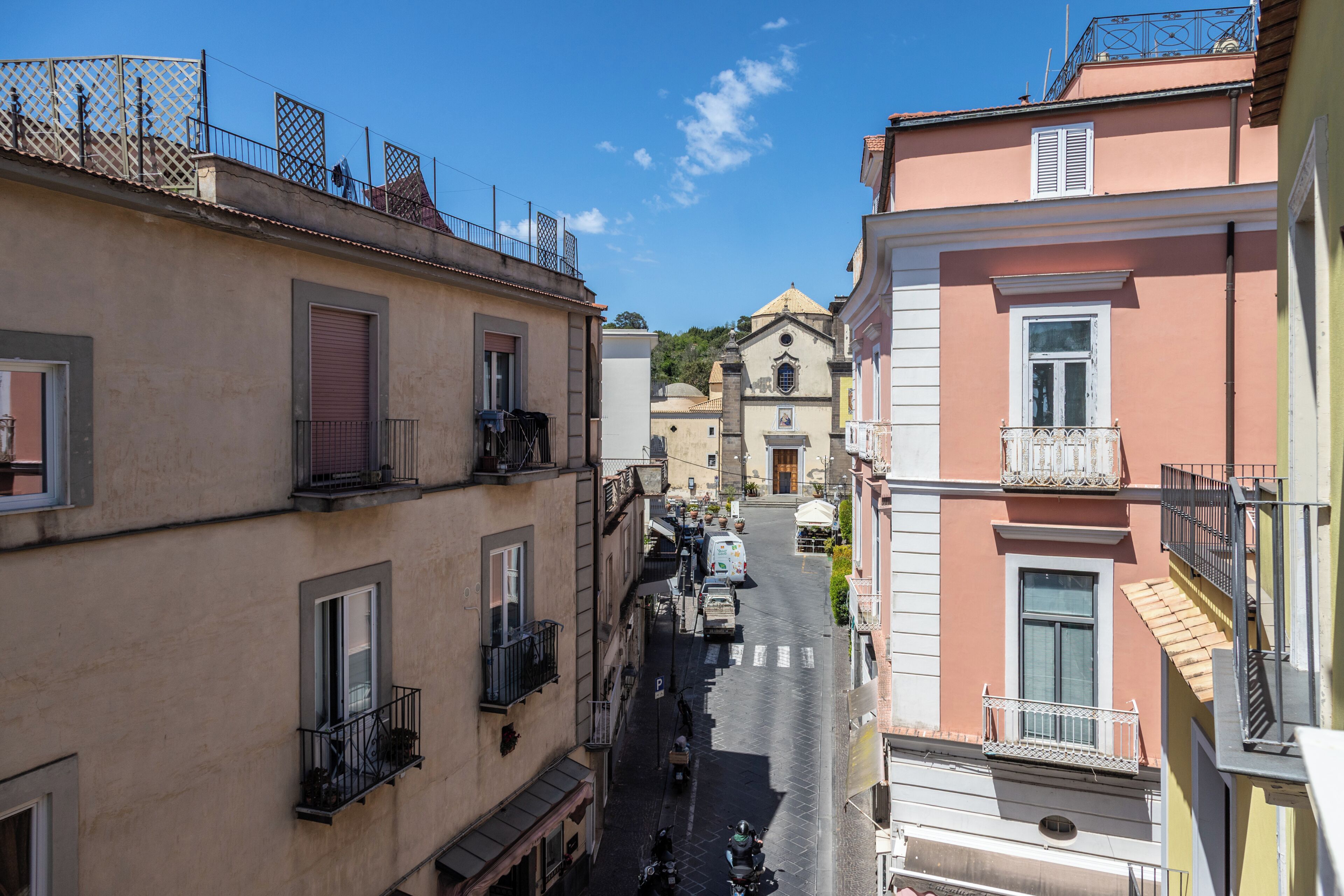 Una calle estrecha bordeada por edificios con balcones, una iglesia al fondo y un cielo azul despejado.
