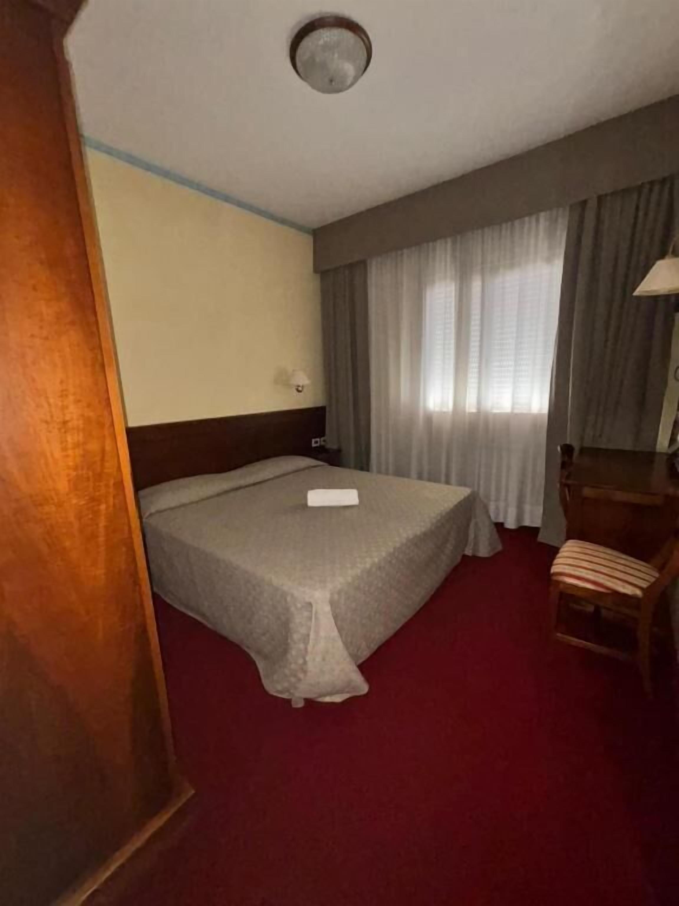 A hotel room with a bed, a desk, a chair, a window with curtains, and a ceiling fan.