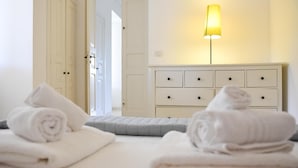 Una habitación de hotel con una cama, una lámpara de noche, un aparador blanco y una puerta que da a otra habitación.