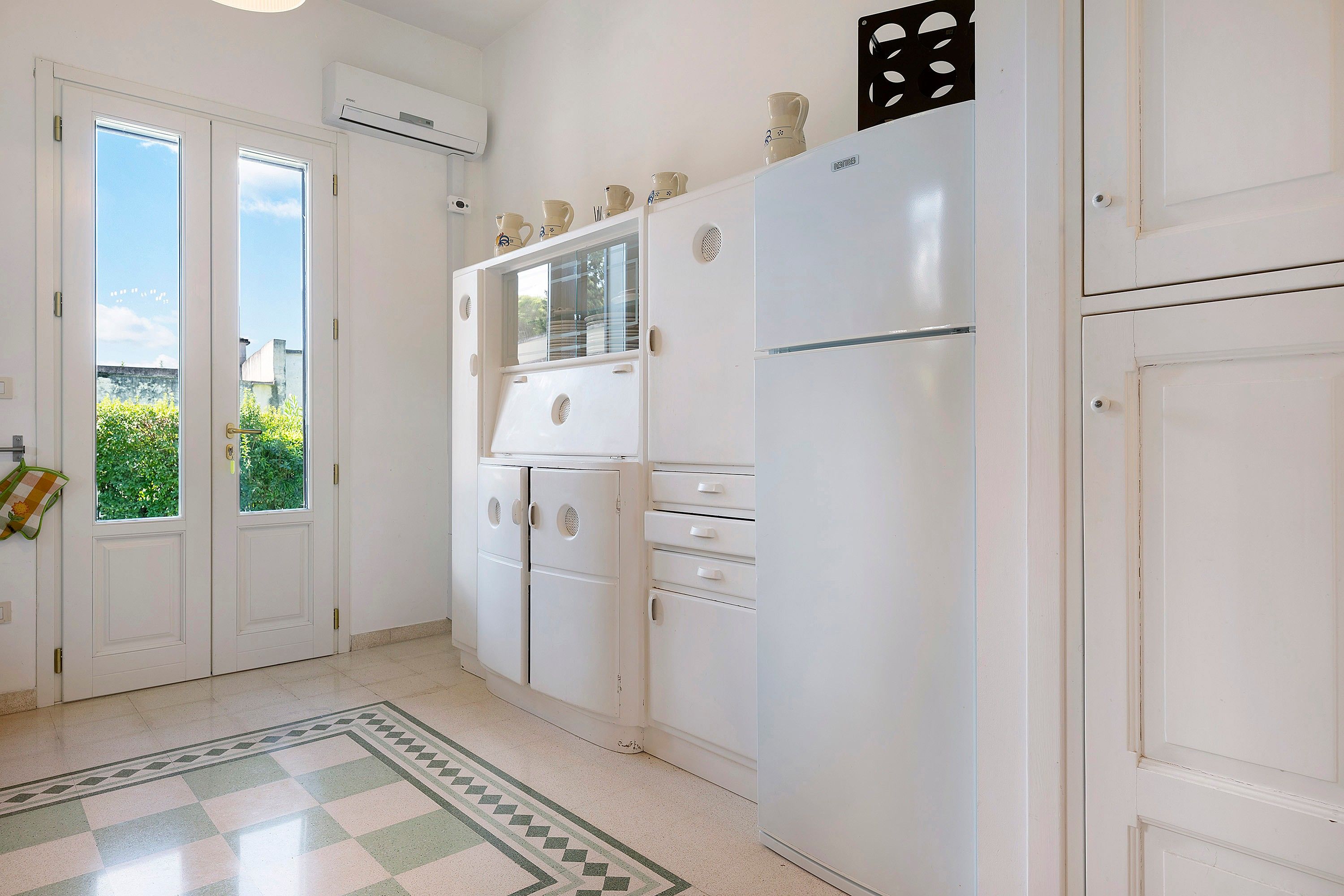 Une cuisine avec des meubles blancs, un réfrigérateur et une fenêtre donnant sur un espace verdoyant.
