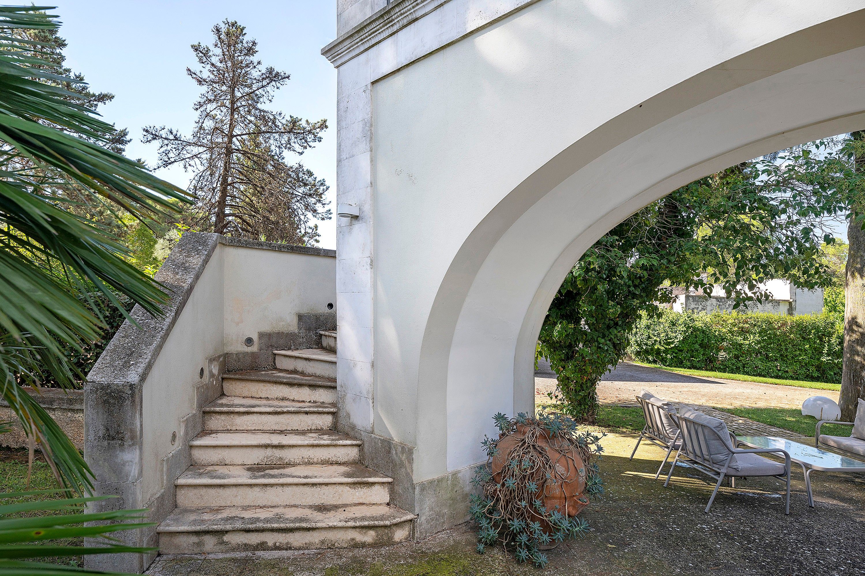 Escadaria de pedra que leva a um arco branco, cercada por vegetação e móveis de jardim.