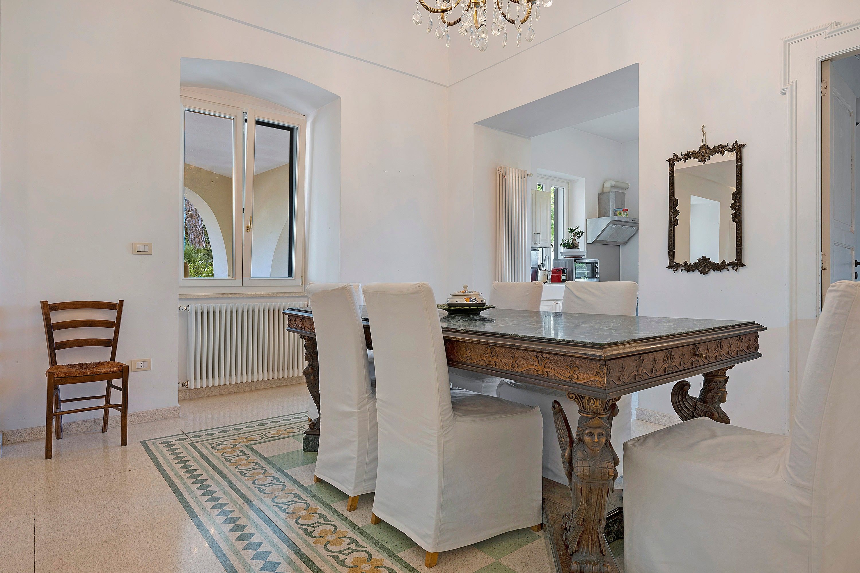 Un espace repas avec une table en marbre, des chaises à revêtement blanc, un lustre et une vue à travers une fenêtre en arc de cercle.