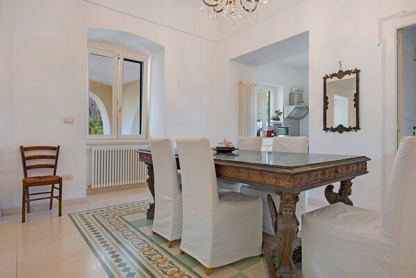 A dining area with a marble table, white upholstered chairs, a chandelier, and a view through an arched window.
