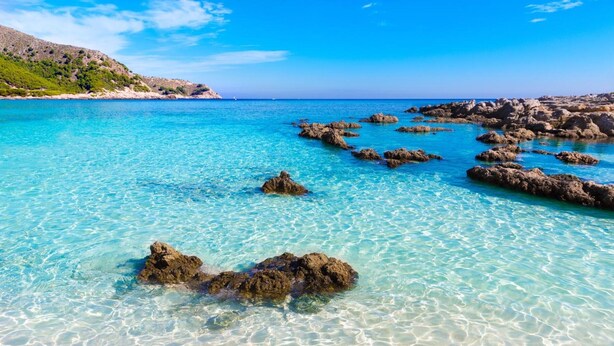 Eine Küstenlandschaft mit klarem türkisblauem Wasser, Felsformationen und einem entfernten Hügel mit vereinzelter Vegetation.