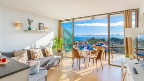 Ein modernes Wohnzimmer mit Sofa, Esstisch und Stühlen, ein Balkon mit Blick und ein großes Fenster.