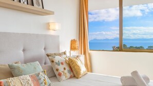 Ein Schlafzimmer mit großem Fenster, einem Bett mit vielen Kissen, einer Nachttischlampe und Blick aufs Meer.