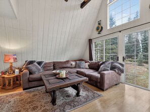 Ein gemütliches Wohnzimmer mit einem Ecksofa, einem Holztisch und einem großen Fenster mit Blick auf Bäume.