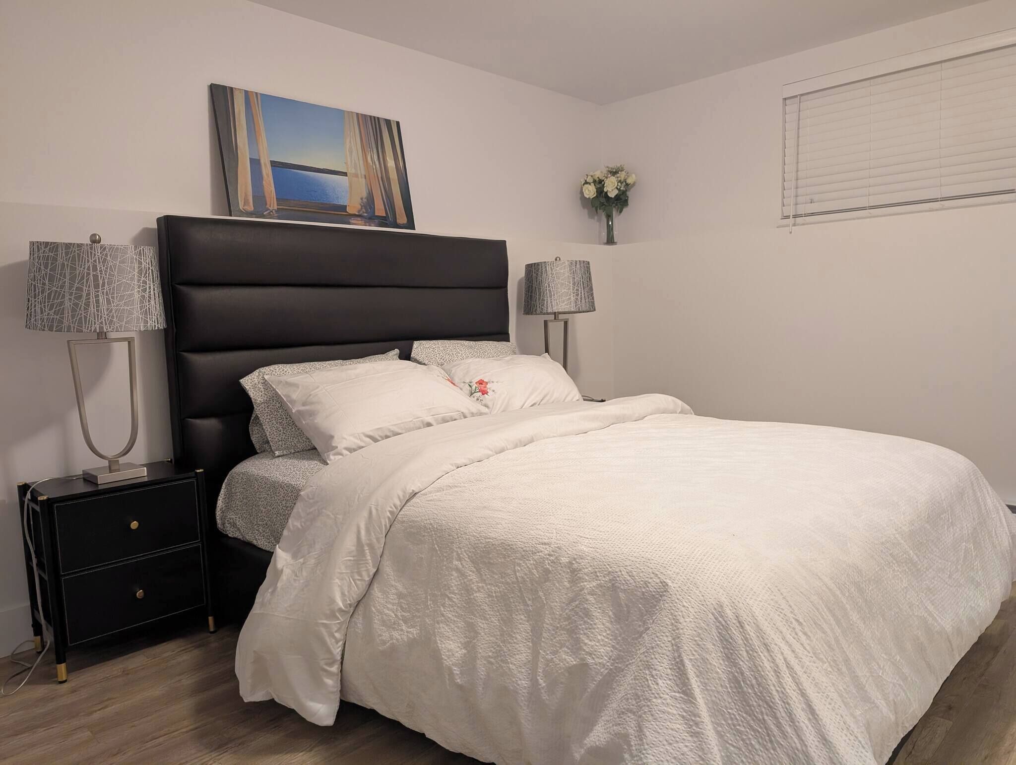 A neatly made bed with a white comforter, two bedside lamps, a black nightstand with drawers, a wall-mounted painting, and a window with blinds.