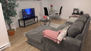 A living room with a grey sofa, a TV, a potted plant, a coffee machine, and a dining area with chairs and a table. - Charming Haven House (Moncton)