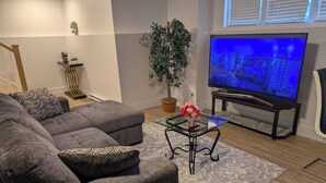 A modern living room with a grey sectional sofa, a glass coffee table, a potted plant, a flat-screen TV, and a staircase. - Charming Haven House (Moncton)