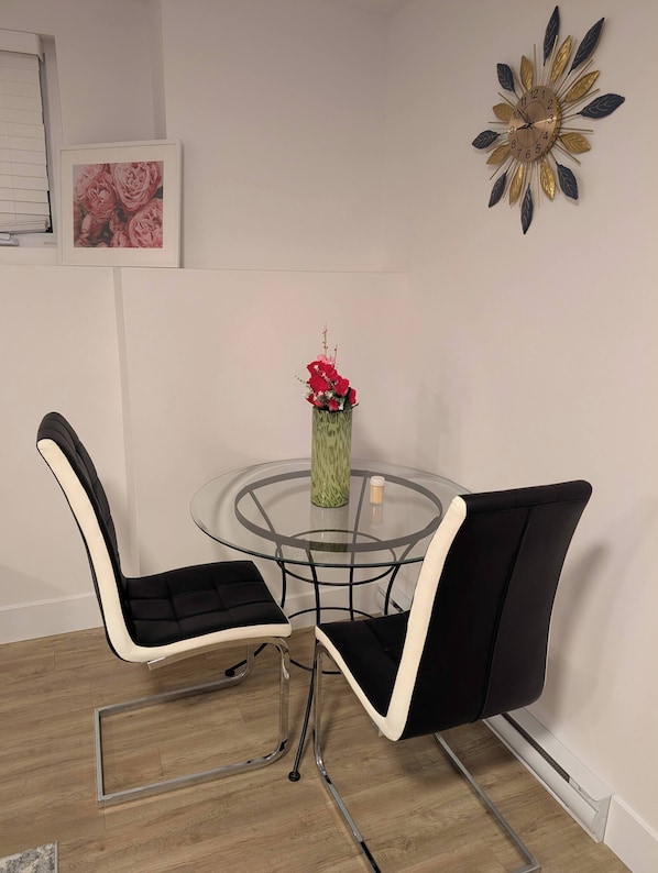 A dining area with a glass table, two black chairs with white cushions, a vase with red flowers, and a wall clock.
