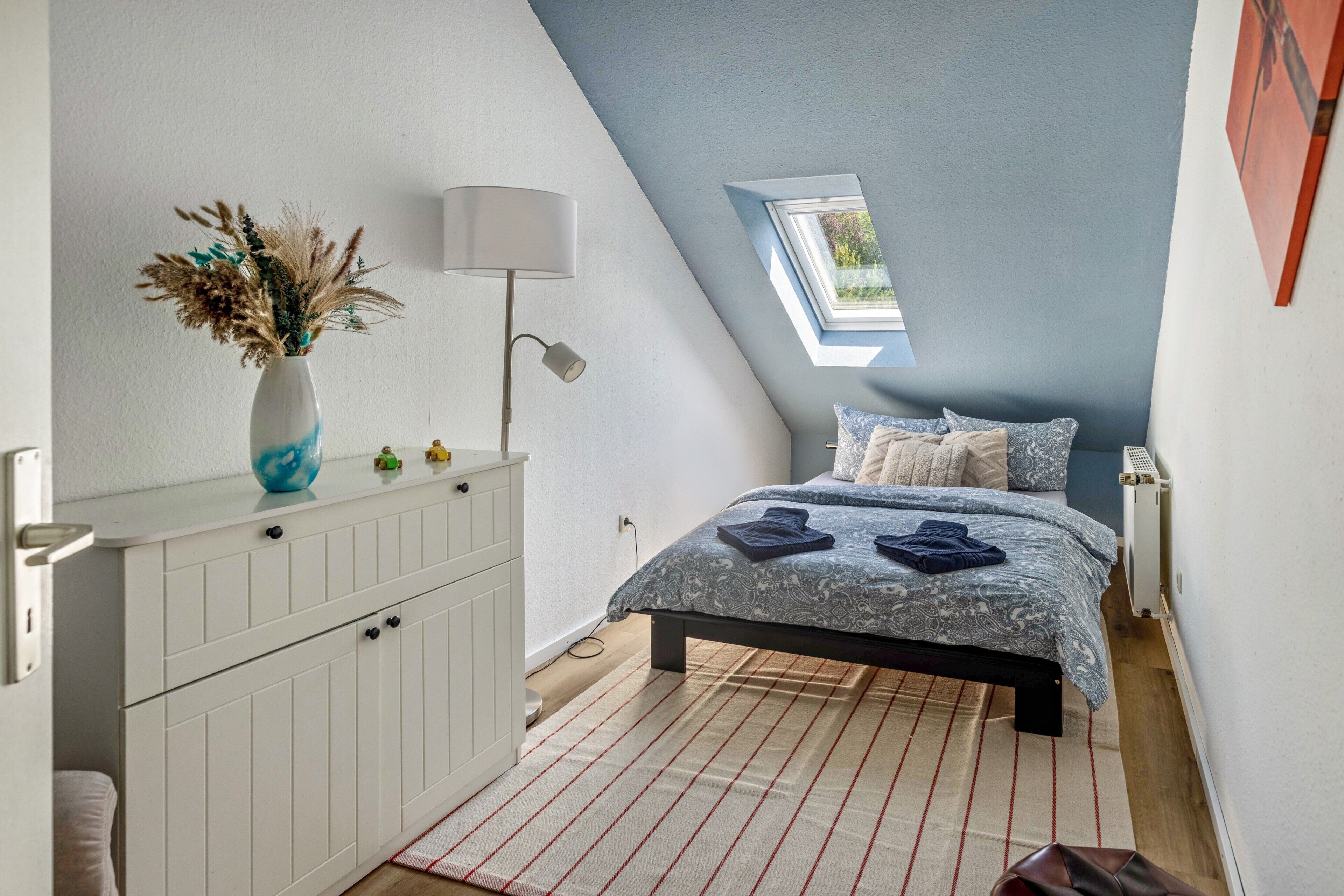 A bedroom with a sloped ceiling, a skylight, a bed with patterned bedding, a bedside table with a lamp, and a dresser.